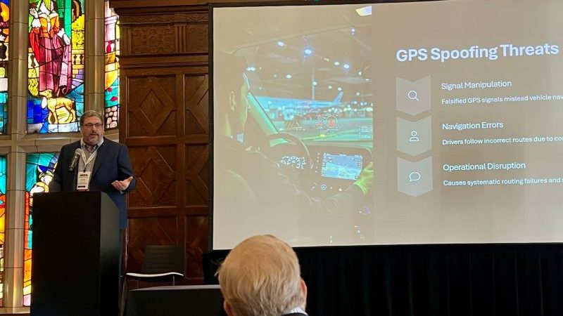 Kevin speaks in front of a projector showing a slide with the title "GPS Spoofing Threats"
